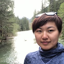 portrait photograph of an East Asian woman with short brown/black hair with a river and forest in the background